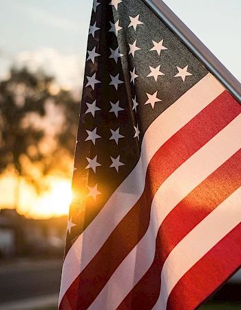 An American flag flapping in the wind at sunset, stars and stripes prominent. The scene feels patriotic and warm.