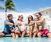 Four friends toast by a pool in bright swimwear, clinking glasses as palm trees and a sunny sky frame the scene.