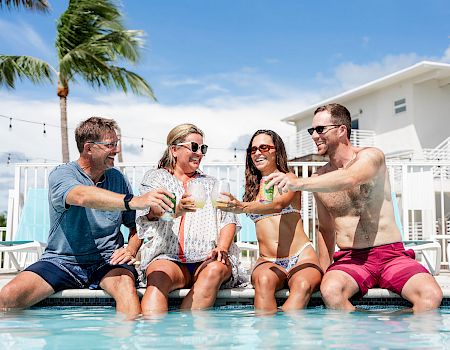 Four friends toast by a pool in bright swimwear, clinking glasses as palm trees and a sunny sky frame the scene.