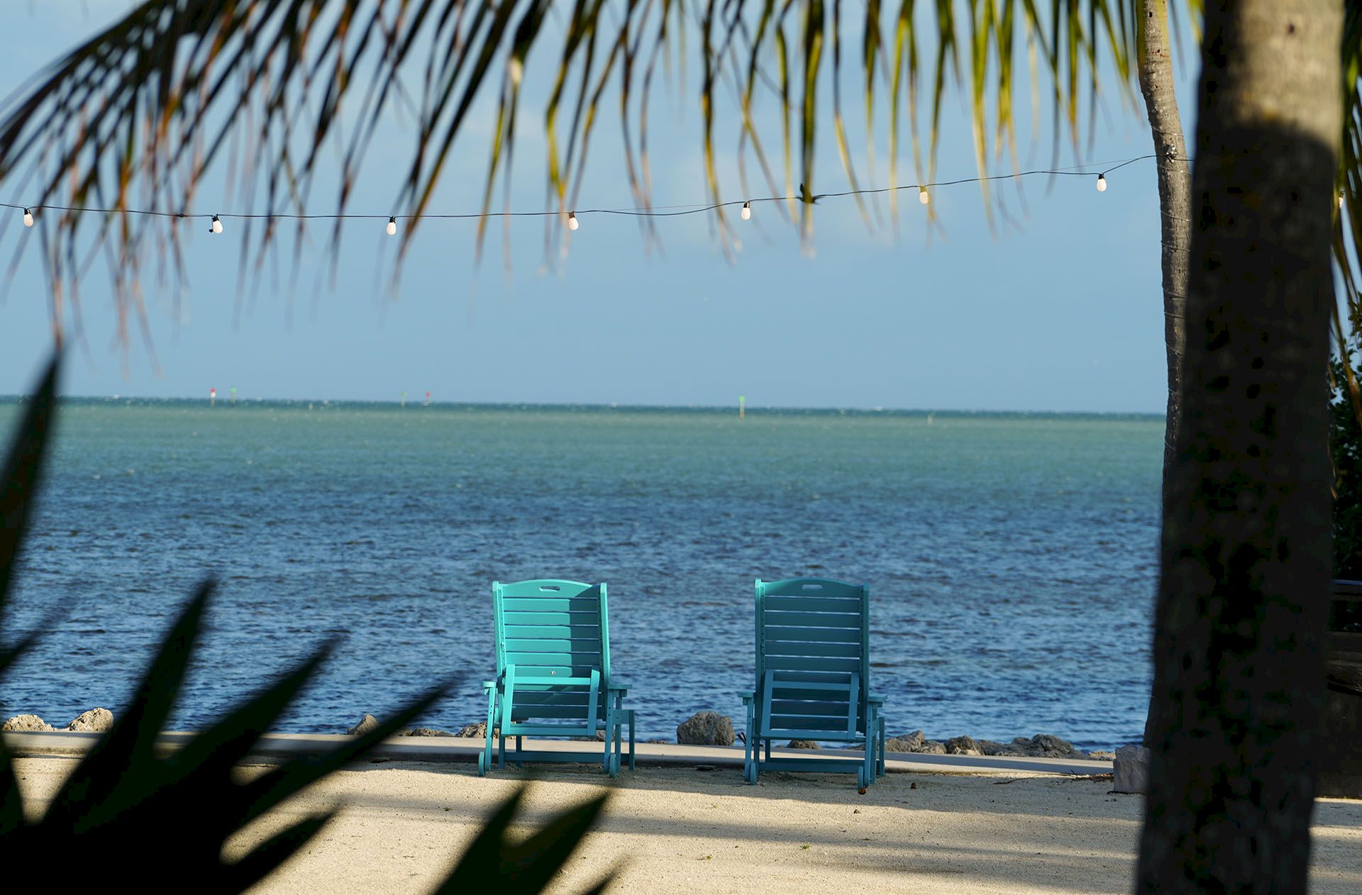 Two teal beach chairs face a calm ocean under palm fronds and hanging string lights.
