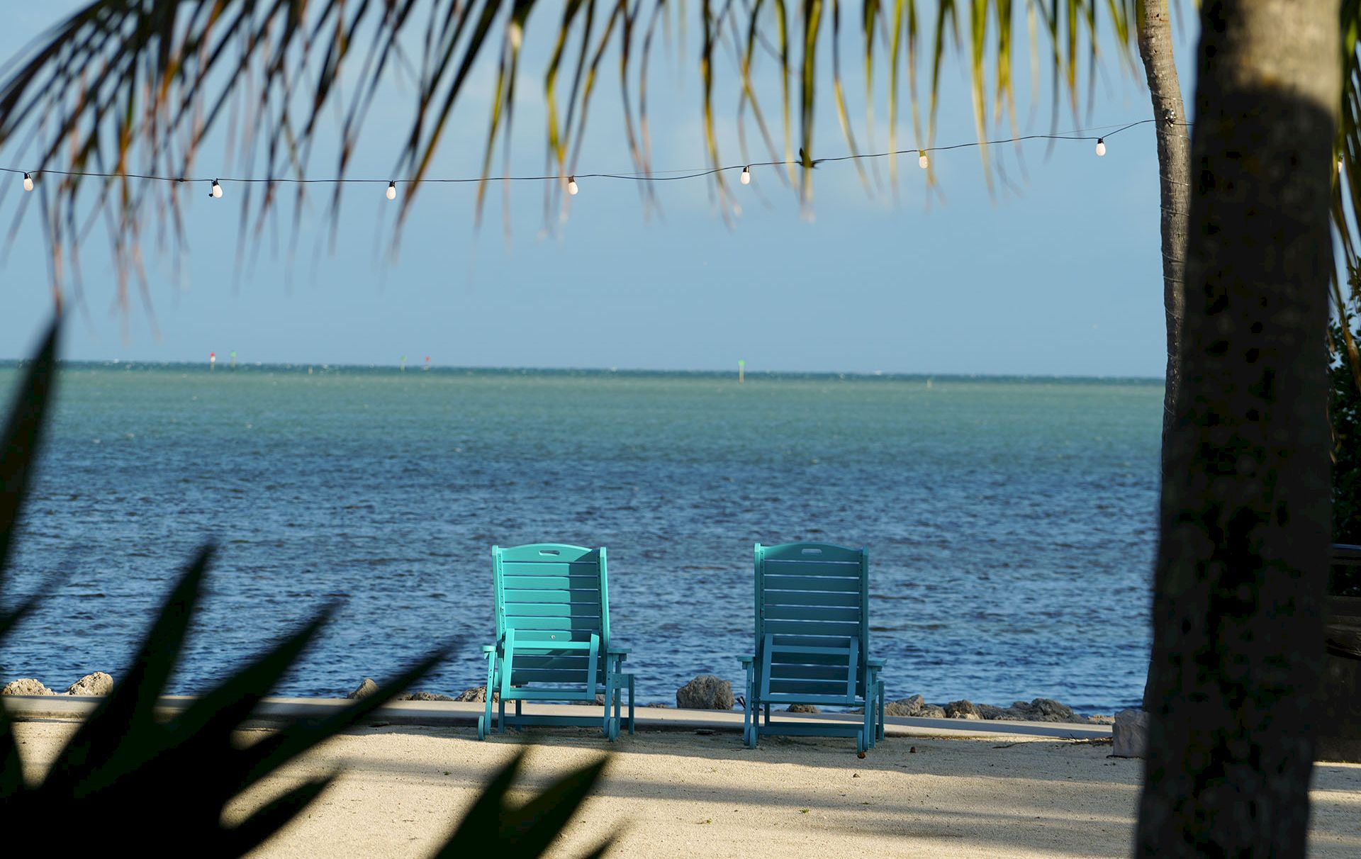 Two teal beach chairs face a calm ocean under palm fronds and hanging string lights.