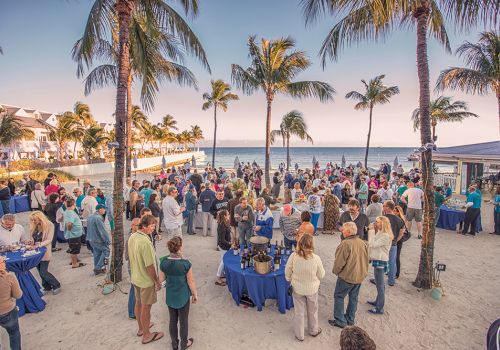Crowd mingles at a seaside cocktail on a sandy beach under palm trees, with tables of drinks and snacks.