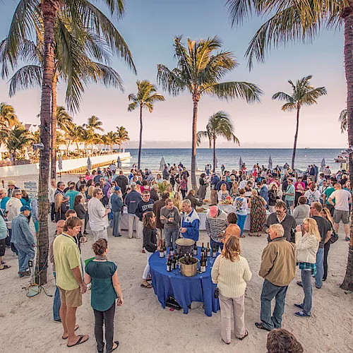 Crowd mingles at a seaside cocktail on a sandy beach under palm trees, with tables of drinks and snacks.