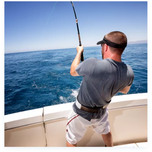 A man stands on a boat holding a fishing rod, casting into the open ocean under a clear blue sky.