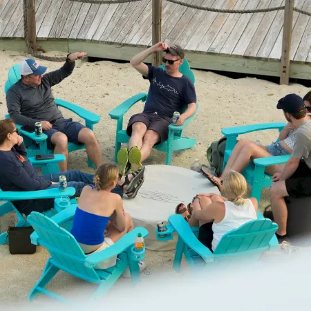 A group of friends relaxing at a backyard picnic, sitting on teal chairs with drinks, snacks, and toys on a table.