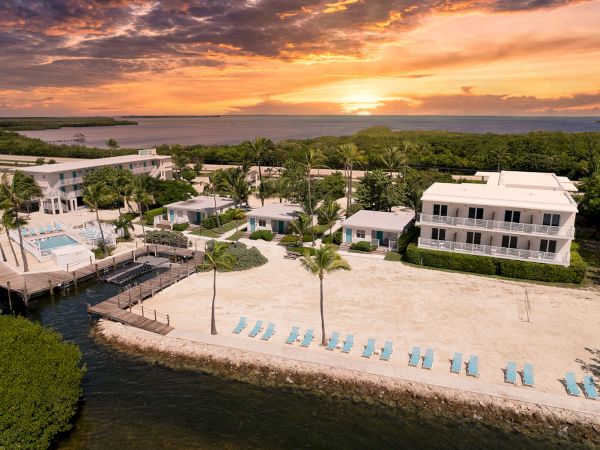 A coastal resort with white buildings, palm trees, docks, and rows of blue loungers facing the ocean at sunset.