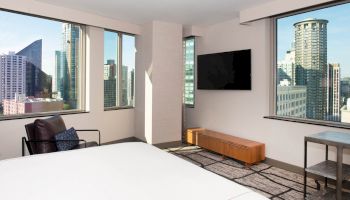 A modern hotel room with a large bed, TV, chair, and city views through tall windows.