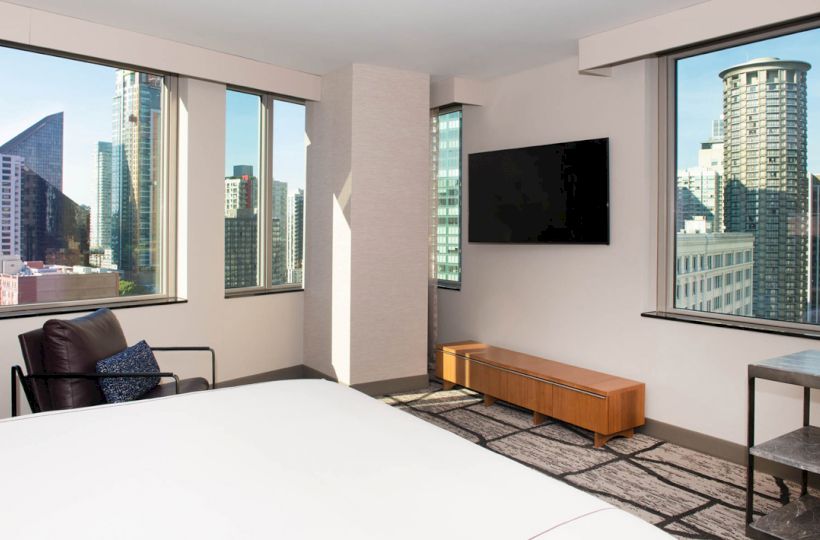 A modern hotel room with a large bed, TV, chair, and city views through tall windows.