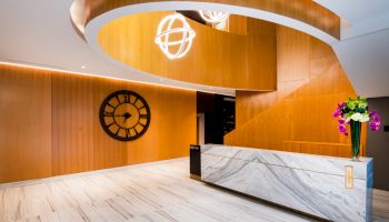 The image shows a modern reception area with wood paneling, a large decorative clock, a marble front desk, and hanging lights.