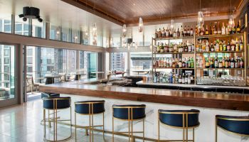 A modern bar with blue and gold stools, a fully stocked shelf with various bottles, and large windows offering a city view, is in the image.