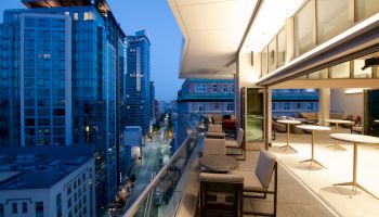 A modern balcony with seating and tables overlooks a city street and surrounding high-rise buildings at twilight.