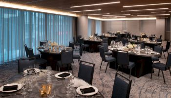 The image shows an elegantly set dining room with multiple round tables, black chairs, silverware, glassware, and floral centerpieces under modern lighting.