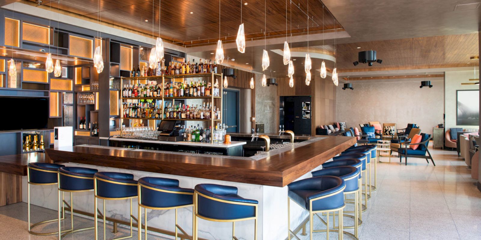 A modern bar with a wide selection of bottles, stylish pendant lights, blue bar stools, and a spacious seating area in the background.