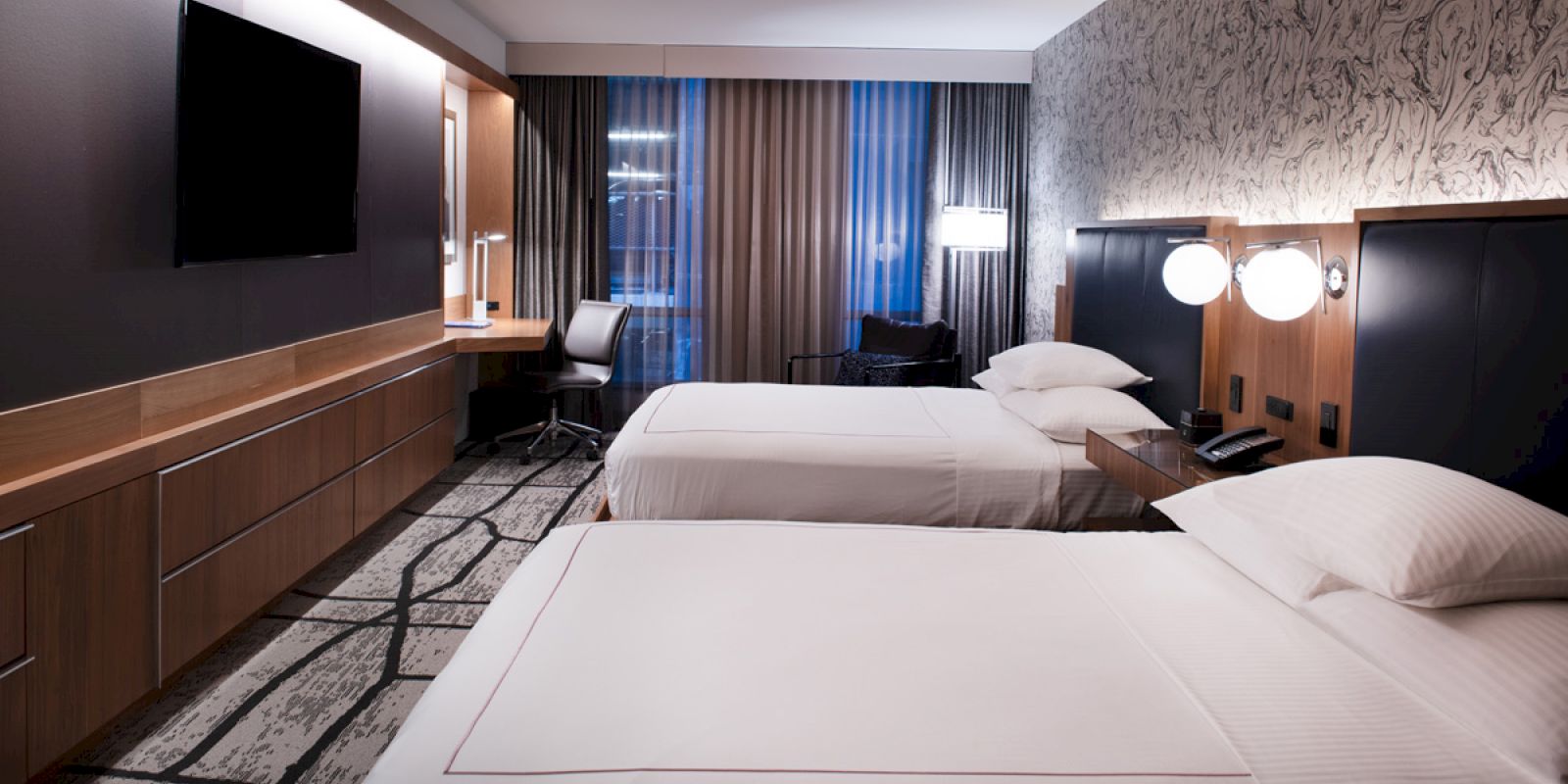 A modern hotel room with two neatly made beds, a desk and chair, large TV, and stylish decor.