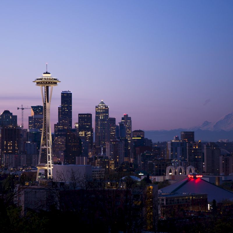 The image shows Seattle's skyline at dusk, featuring the Space Needle and Mount Rainier in the background under a purple-blue sky.