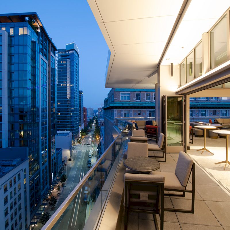 A modern cityscape at dusk with high-rise buildings, a street below, and a sleek, furnished balcony extending from a contemporary building.