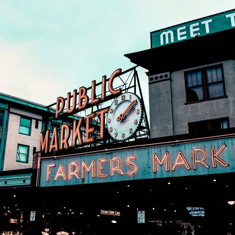 The image shows an exterior view of a "Public Market" with a "Farmers Market" sign below it, featuring a clock and adjacent buildings in the background.