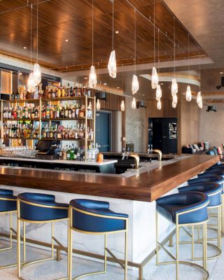 A modern bar with a central counter, blue stools, hanging lights, and a well-stocked shelf of alcohol bottles, accompanied by lounge seating.