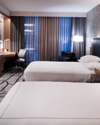 A modern hotel room with two beds, a wall-mounted TV, a work desk, and contemporary decor, including patterned wallpaper and a large window.