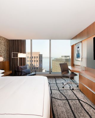 A modern hotel room features a large bed, a desk with a chair, a wall-mounted TV, and a window offering a cityscape view.