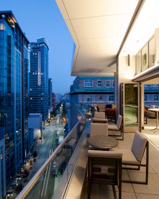 A modern balcony with tables and chairs overlooks a cityscape of tall buildings at dusk, featuring illuminated streets and a serene ambiance.