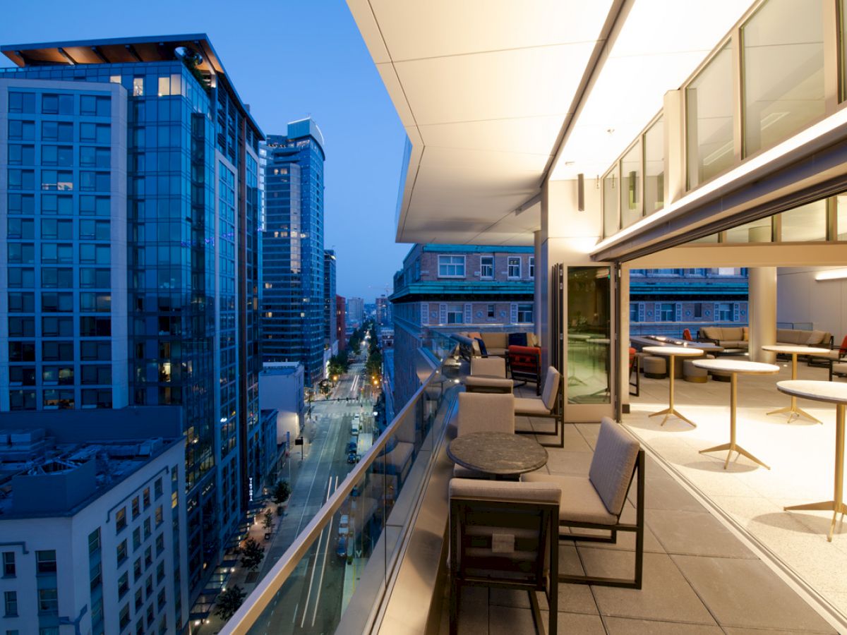 A modern urban balcony with tables and chairs overlooks a city street lined with high-rise buildings, captured at dusk.