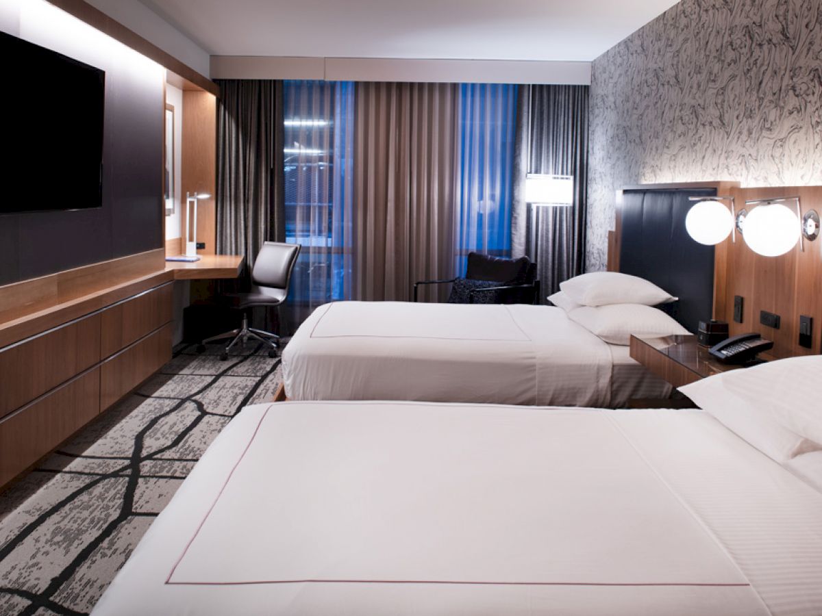 This image shows a modern hotel room with two beds, a wall-mounted TV, a desk with a chair, patterned wallpaper, and stylish lighting.