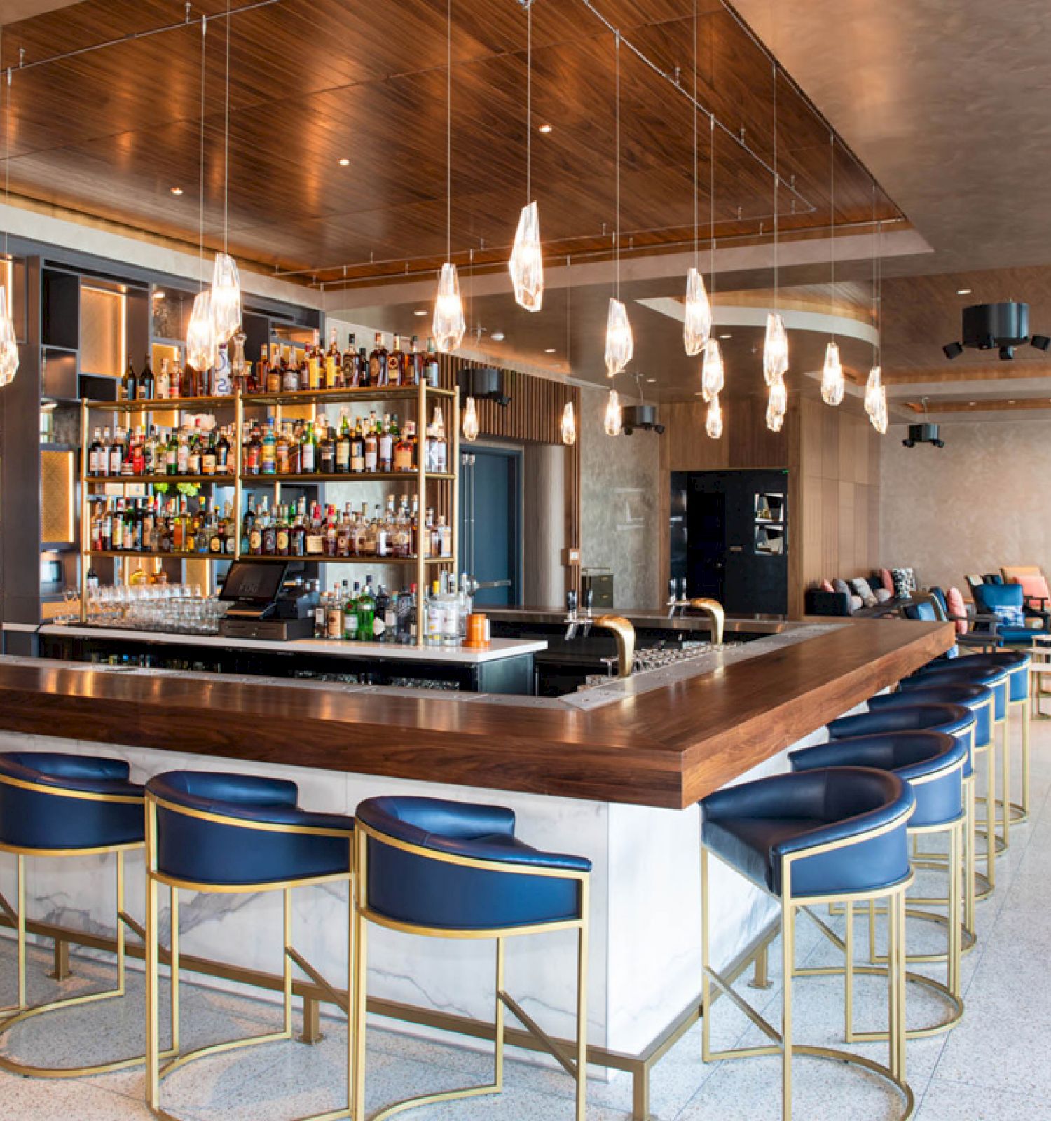 This image shows a modern bar with a central counter, blue chairs, hanging lights, and a variety of bottles displayed. There is seating in the background.