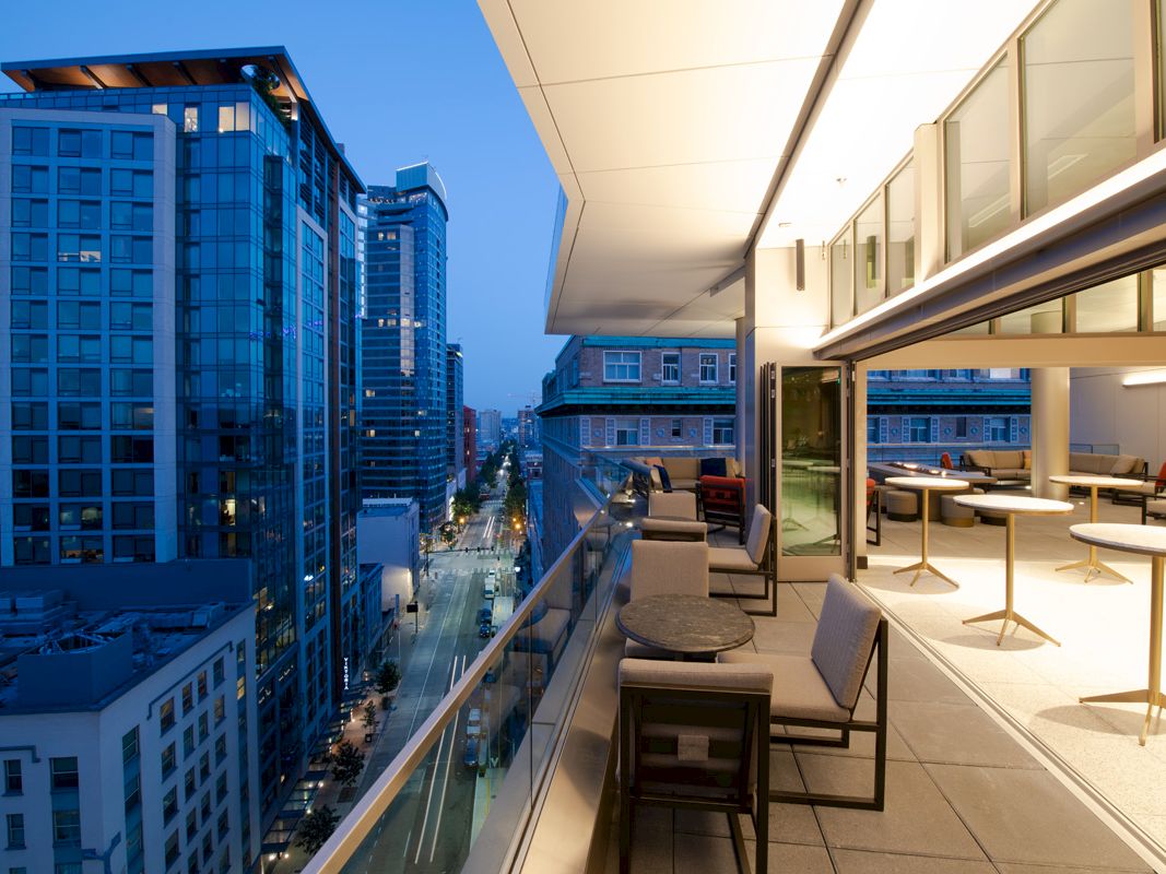 The image depicts a modern cityscape with high-rise buildings and a balcony overlooking a street, furnished with tables and chairs. The sentence ends.