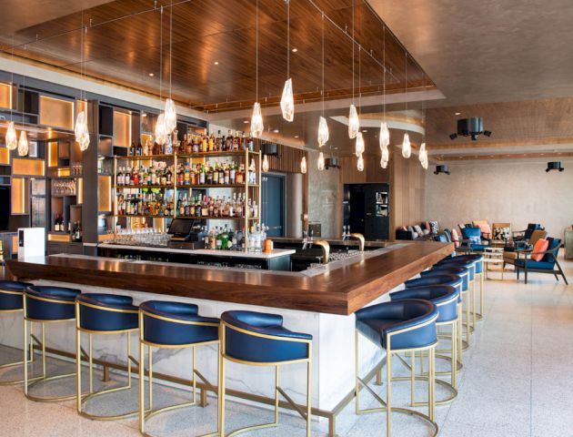 The image shows a modern bar with a sleek design, featuring a long wooden counter, blue bar stools, a variety of bottles, and hanging light fixtures.