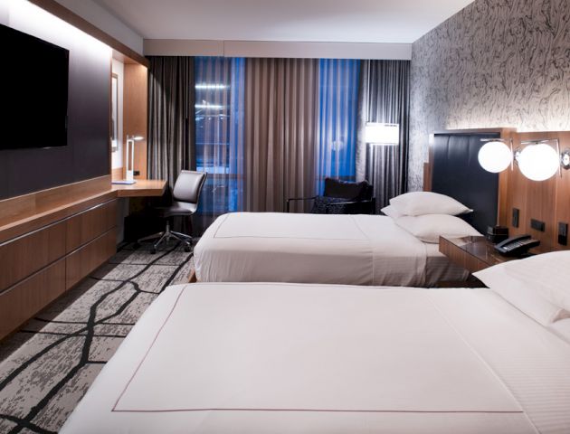 A modern hotel room with two double beds, a flat-screen TV, a desk with a chair, and stylish decor.