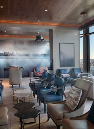 The image shows a modern lounge area with comfortable seating, tables, and large windows offering a scenic view of a waterfront and a Ferris wheel.