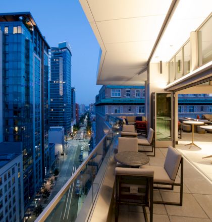 A cityscape at dusk featuring high-rise buildings and a modern balcony with chairs and tables, providing a view of the street below.