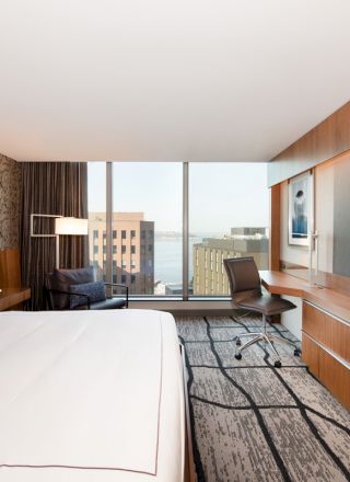 A modern hotel room with a large bed, sleek wooden desk, mounted TV, lounge chair by a window, and a view of buildings and water outside.