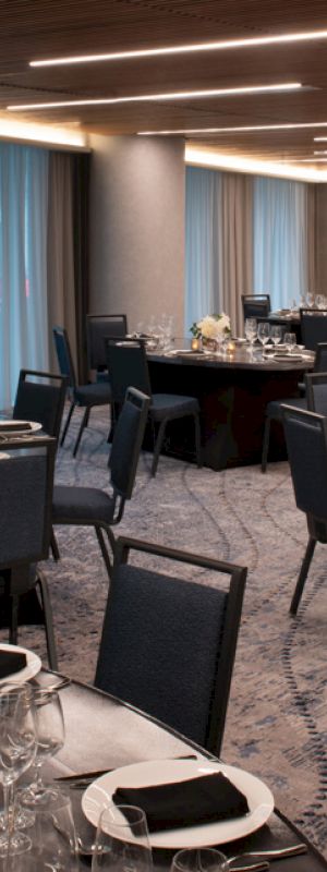 A banquet hall set up with round tables, elegant place settings, and floral centerpieces. The room features modern lighting and floor-to-ceiling windows.