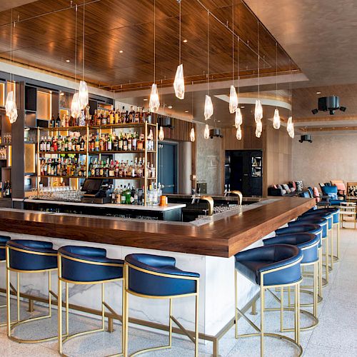 A modern bar with blue chairs, a well-stocked liquor shelf, and pendant lights above the counter. There is seating in the background.