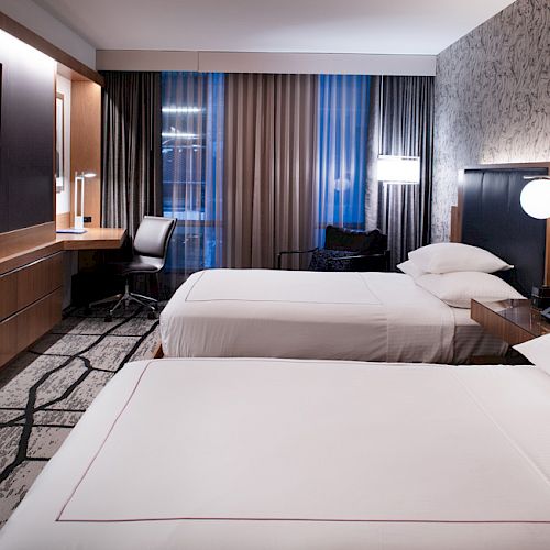 A modern hotel room with two neatly made beds, a wall-mounted TV, a desk, and large windows with curtains.