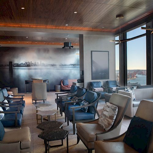 A modern lounge with leather chairs, a cityscape artwork, large windows offering waterfront views, and contemporary lighting fixtures, ending the sentence.