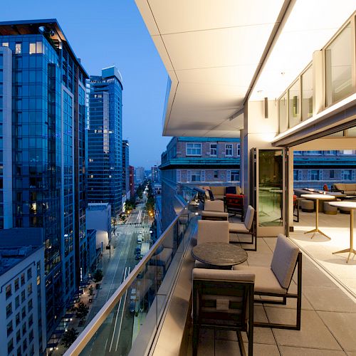 A modern cityscape at dusk with tall buildings, a street below, and a luxurious rooftop patio featuring tables, chairs, and soft, ambient lighting.