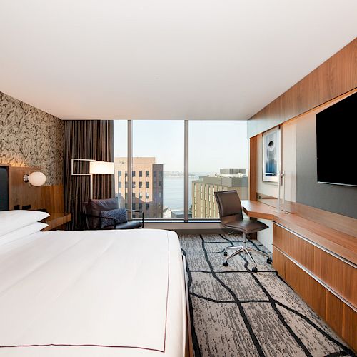 A modern hotel room with a large bed, wall-mounted TV, desk, and a view of buildings and water through floor-to-ceiling windows.