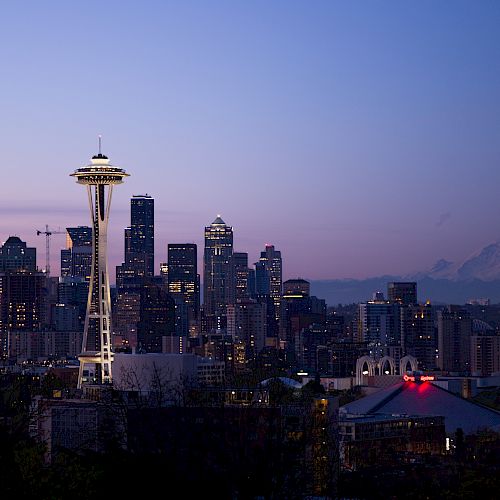 A cityscape of Seattle at dusk, featuring the Space Needle, downtown skyscrapers, and Mount Rainier in the background, illuminated by evening lights.