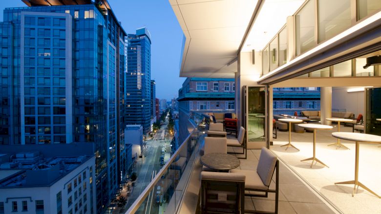 A modern cityscape at dusk features high-rise buildings alongside a spacious balcony with seating and tables, overlooking a busy street.