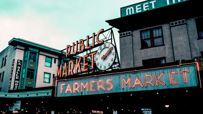 The image shows the iconic neon sign of a public farmers market with a clock, against a backdrop of buildings and a partly clouded sky.