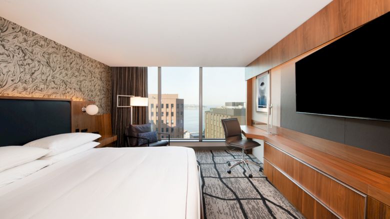 A modern hotel room with a large bed, a wall-mounted TV, a desk with a chair, and a seating area by floor-to-ceiling windows overlooking a cityscape.