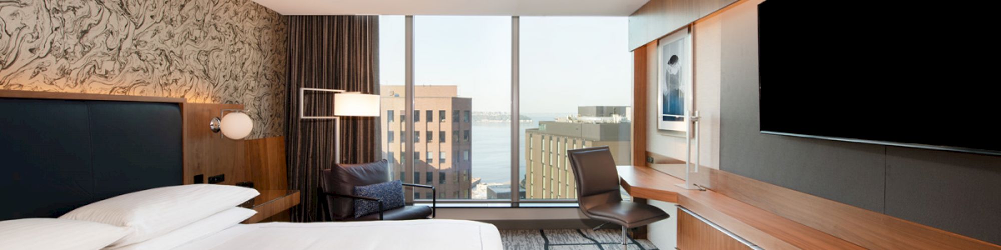 This image shows a modern hotel room with a large bed, a desk, a flat-screen TV, and a view of buildings and water through the window.