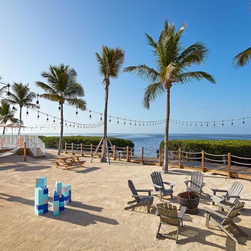 A beachside setting with palm trees, string lights, Adirondack chairs around a fire pit, and a view of the ocean under a clear blue sky.