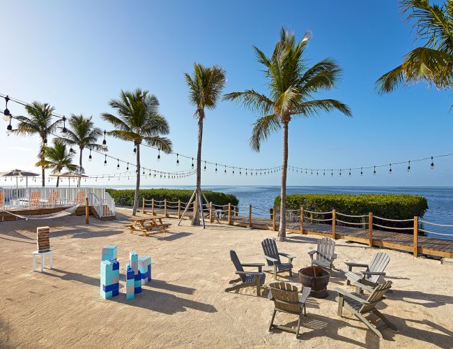 A beachside setting with palm trees, string lights, Adirondack chairs around a fire pit, and a view of the ocean under a clear blue sky.