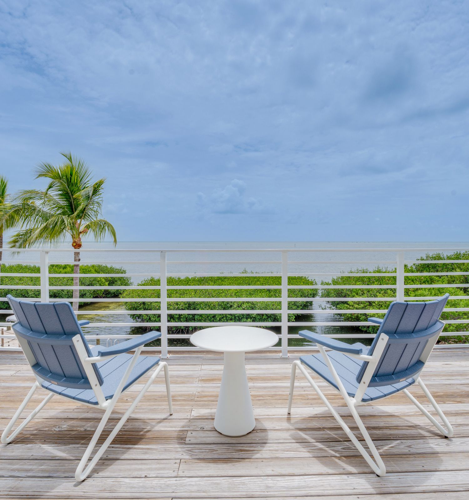 Two chairs and a small table on a wooden deck overlooking the ocean with palm trees in the background under a partly cloudy sky.