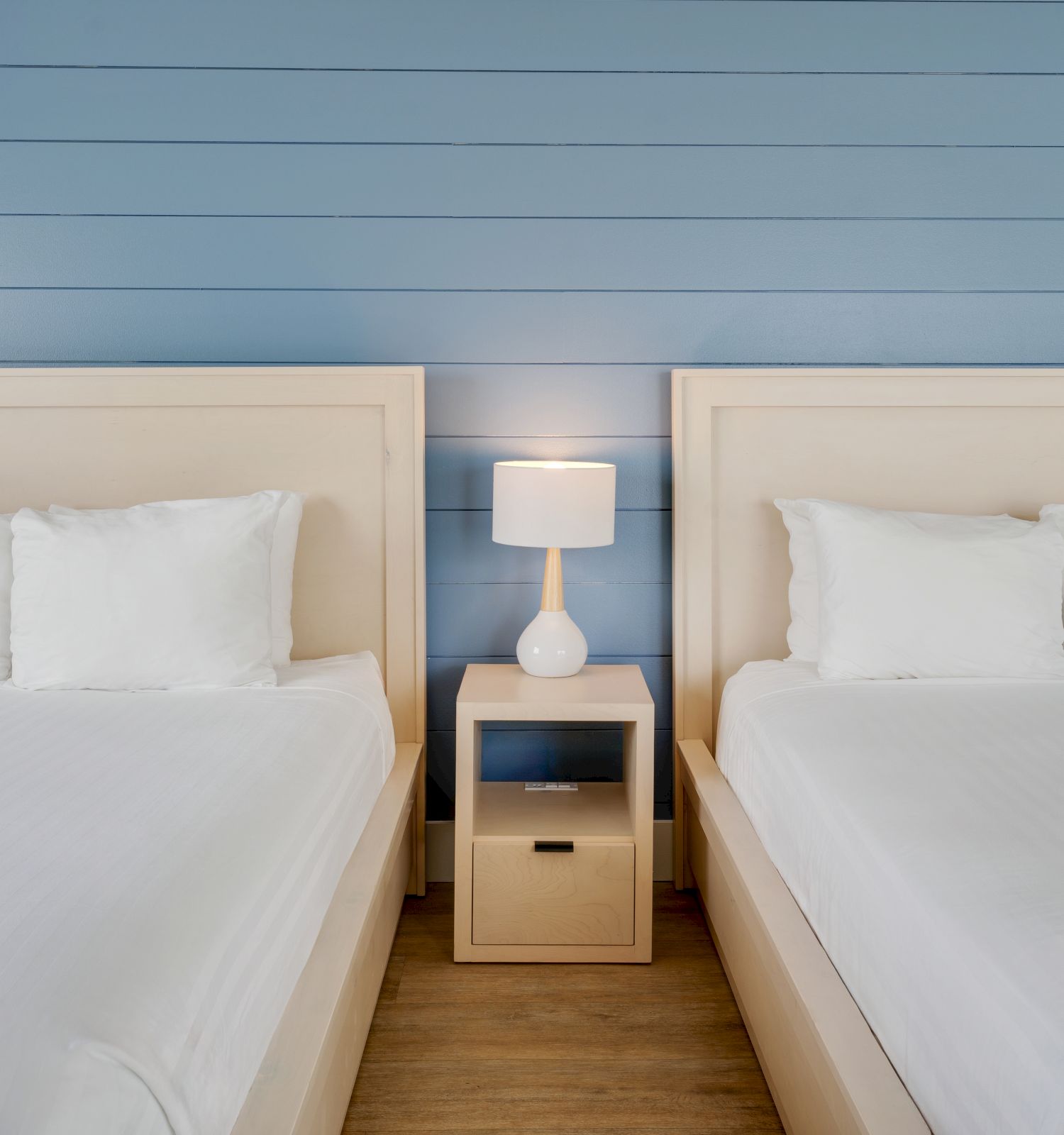 Two neatly made beds with white linens are placed on either side of a nightstand with a lamp, against a blue wall background.