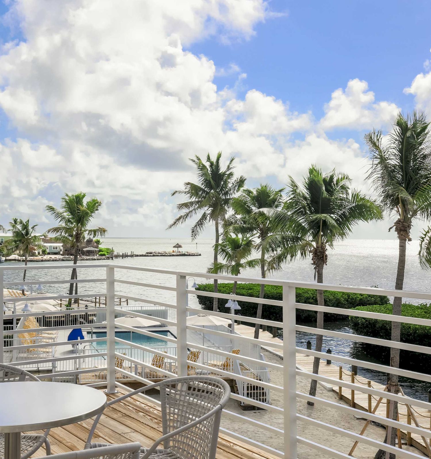 A patio with a table and chairs overlooking a scenic ocean view with palm trees, blue skies, and fluffy clouds.